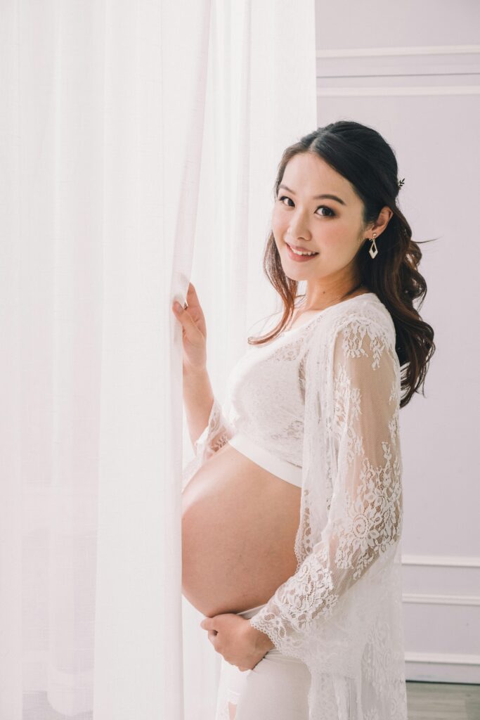NIEPŁODNOŚĆ CIĄŻA Happy young Asian pregnant female in white wear standing near curtain and looking at camera