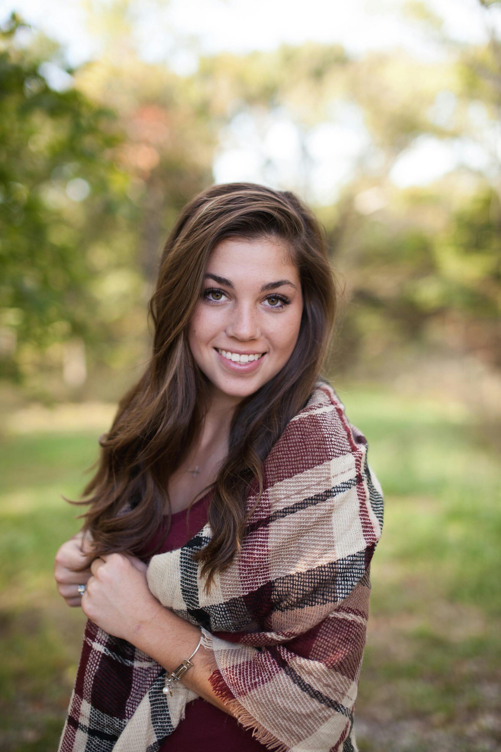 A woman smiling outdoors with a cozy plaid shawl, surrounded by nature in a sunny park.