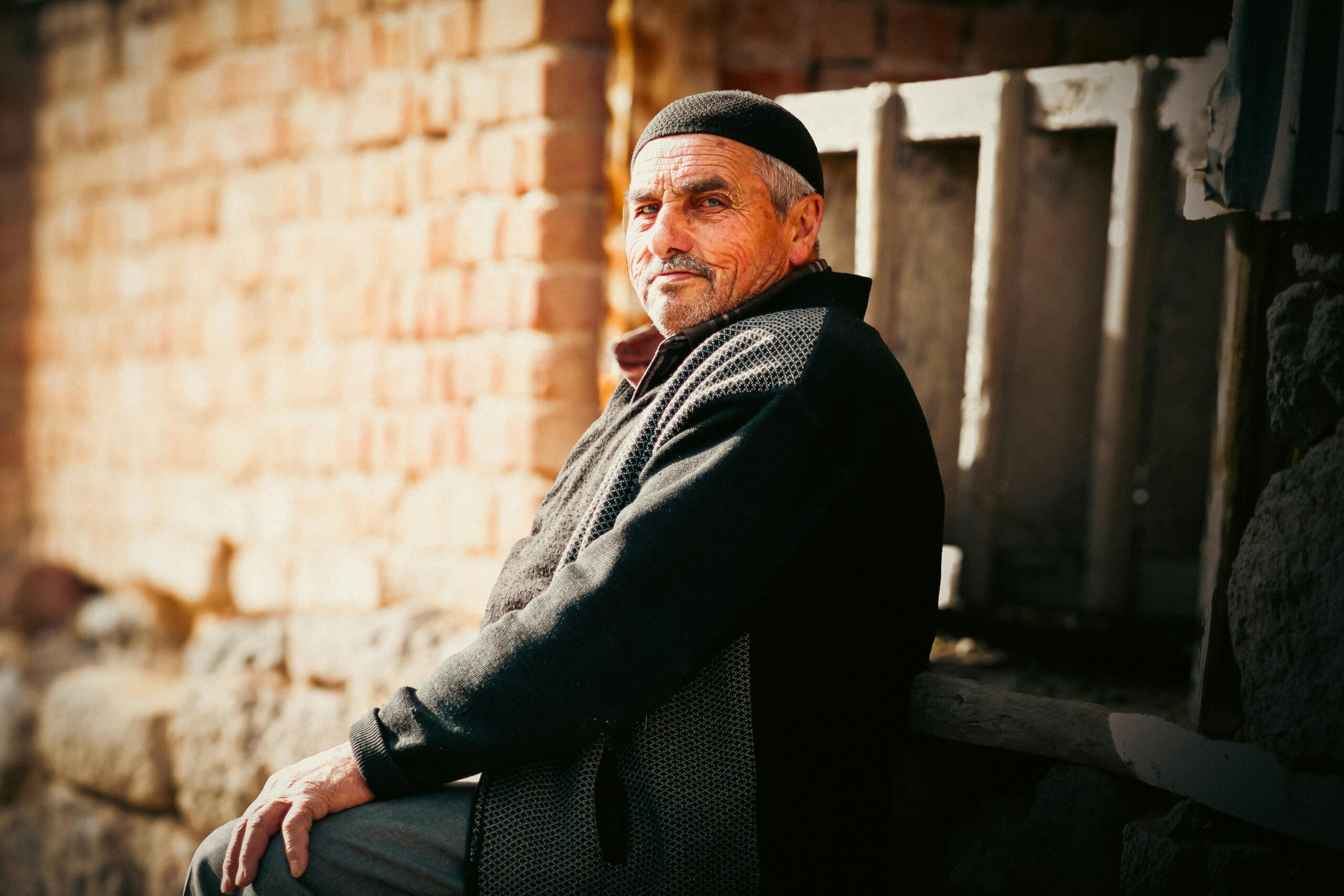 Portrait of an elderly man sitting outdoors against a sunlit brick wall in casual attire.